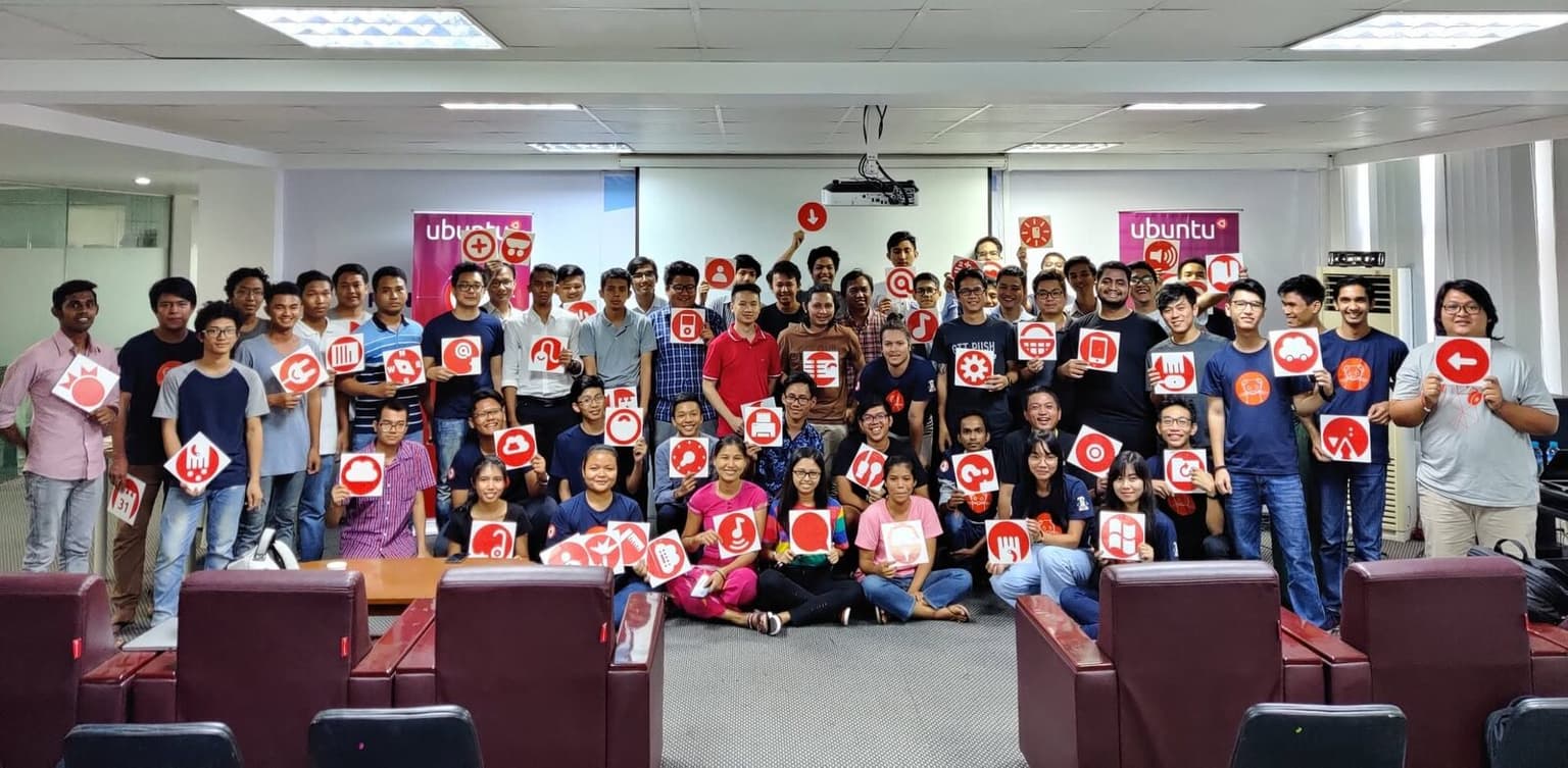 Around sixty members of the Ubuntu Myanmar community gathered in a meeting room, most holding up red-and-white Suru icon cards, with Ubuntu banners on either side.